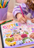 Child holding a pink pencil over the Cuties colouring book with colourful cute characters on the cover.